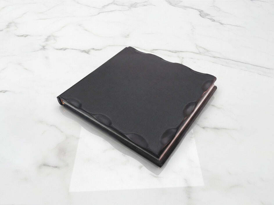 Fotobook clásico rústico Mi Album en color negro, cerrado y apoyado sobre superficie de mármol blanco, con diseño ondulado artesanal y acabado elegante de alta calidad.