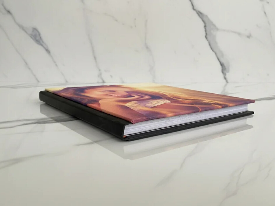 Fotobook Layflat Mi Album con portada en canvas visto en diagonal tipo flat lay, lomo negro en pro-piel y acabado profesional sobre mármol.